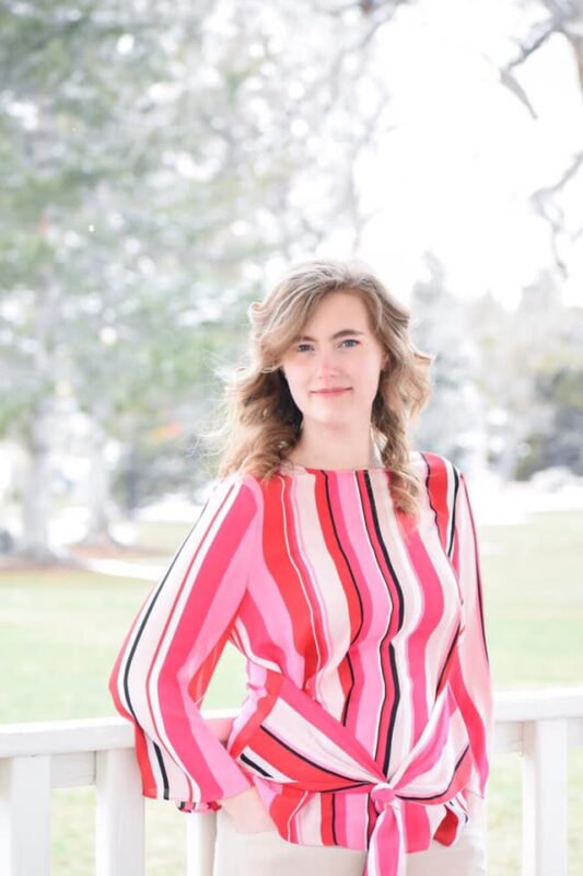 Micayla Bellamy, a caucasian woman with long hair and a neutral, pleasant smile stands casually-dressed in a brightly-lit outdoor setting.